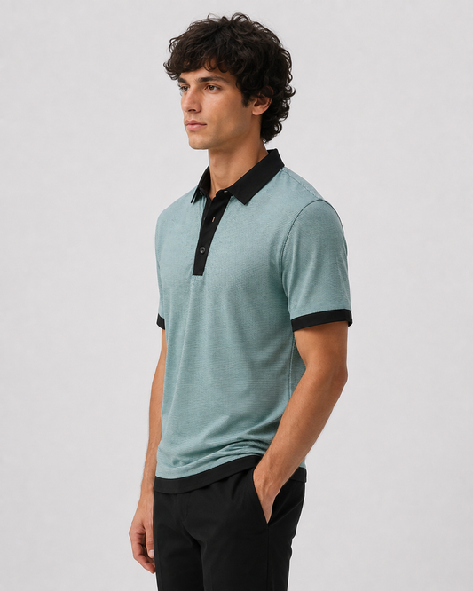 A man wearing a short-sleeve polo shirt in a light teal color with a black collar and black trim on the placket and cuffs, paired with black trousers, photographed against a neutral light gray background.