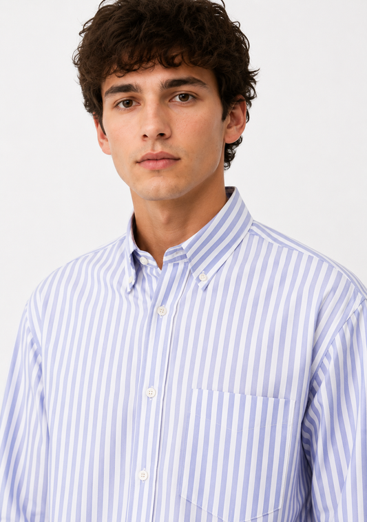A young man with curly dark hair wearing a light blue and white vertically striped button-down shirt, photographed against a clean white studio background.