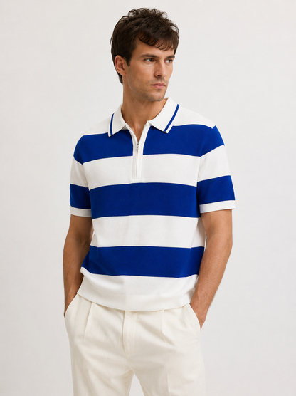 A man wearing a blue and white horizontally striped short-sleeve polo shirt with cream trousers, standing against a plain light gray studio background.