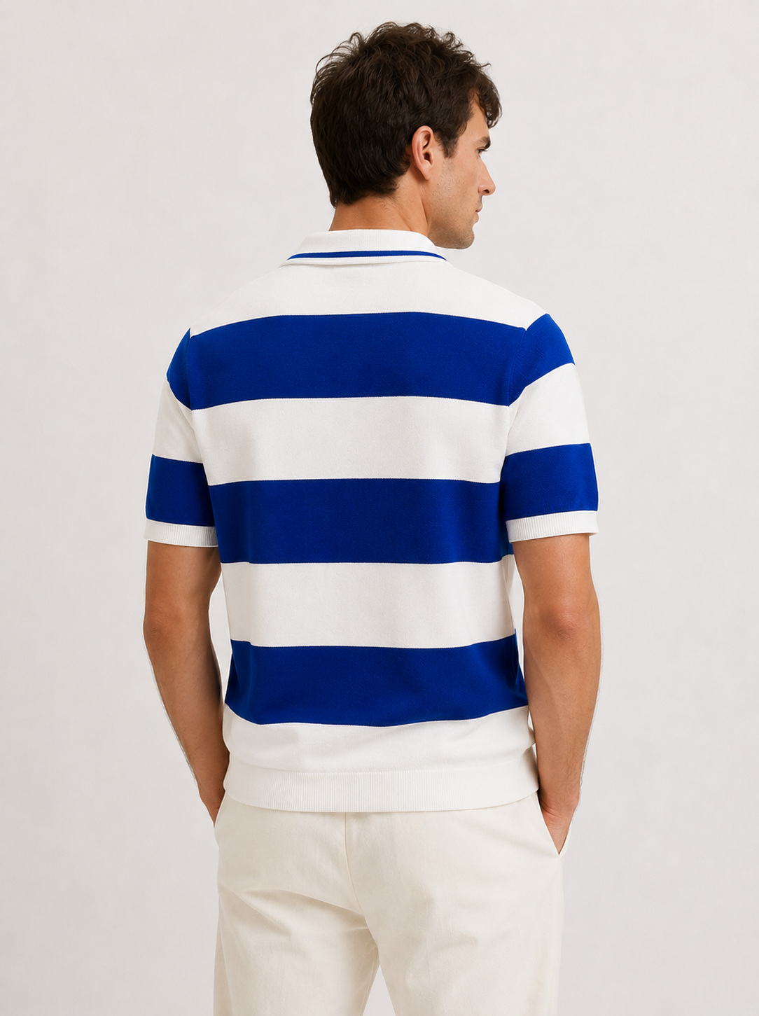 A man viewed from the back wearing a short-sleeve polo shirt with bold horizontal blue and white stripes, paired with off-white trousers, against a plain light gray studio background.