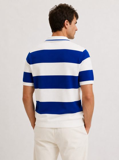 A man viewed from the back wearing a short-sleeve polo shirt with bold horizontal blue and white stripes, paired with off-white trousers, against a plain light gray studio background.
