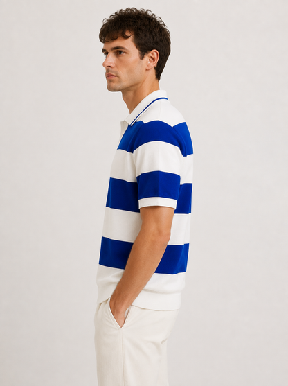 A man wearing a blue and white horizontally striped short-sleeve polo shirt, paired with off-white trousers, standing against a plain light gray studio background.