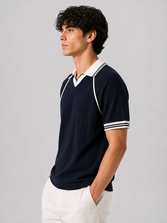 A man wearing a navy blue ribbed short-sleeve polo shirt with white contrast trim on the collar, shoulders, and sleeve cuffs, paired with white trousers, photographed against a neutral gray studio background.